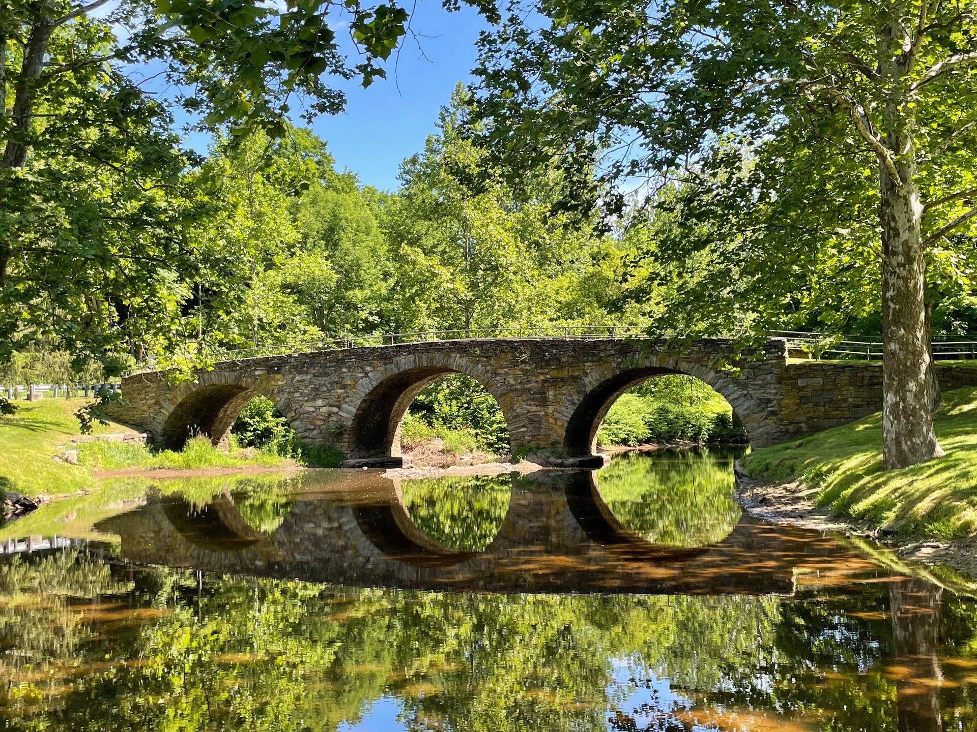 Eine steinerne Brücke spannt sich über einen Fluss. Die Ufer sind mit Gras und Bäumen bewachsen. Im Fluss spiegelt sich die Brücke.