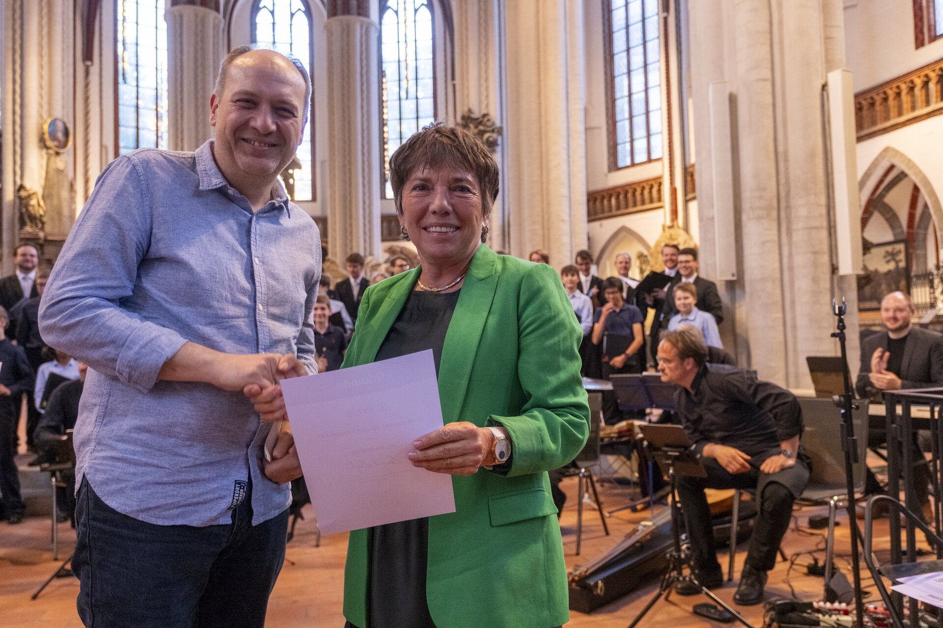 Ein Mann in blauem Hemd reicht einer Frau in grünem Blazer die Hand.