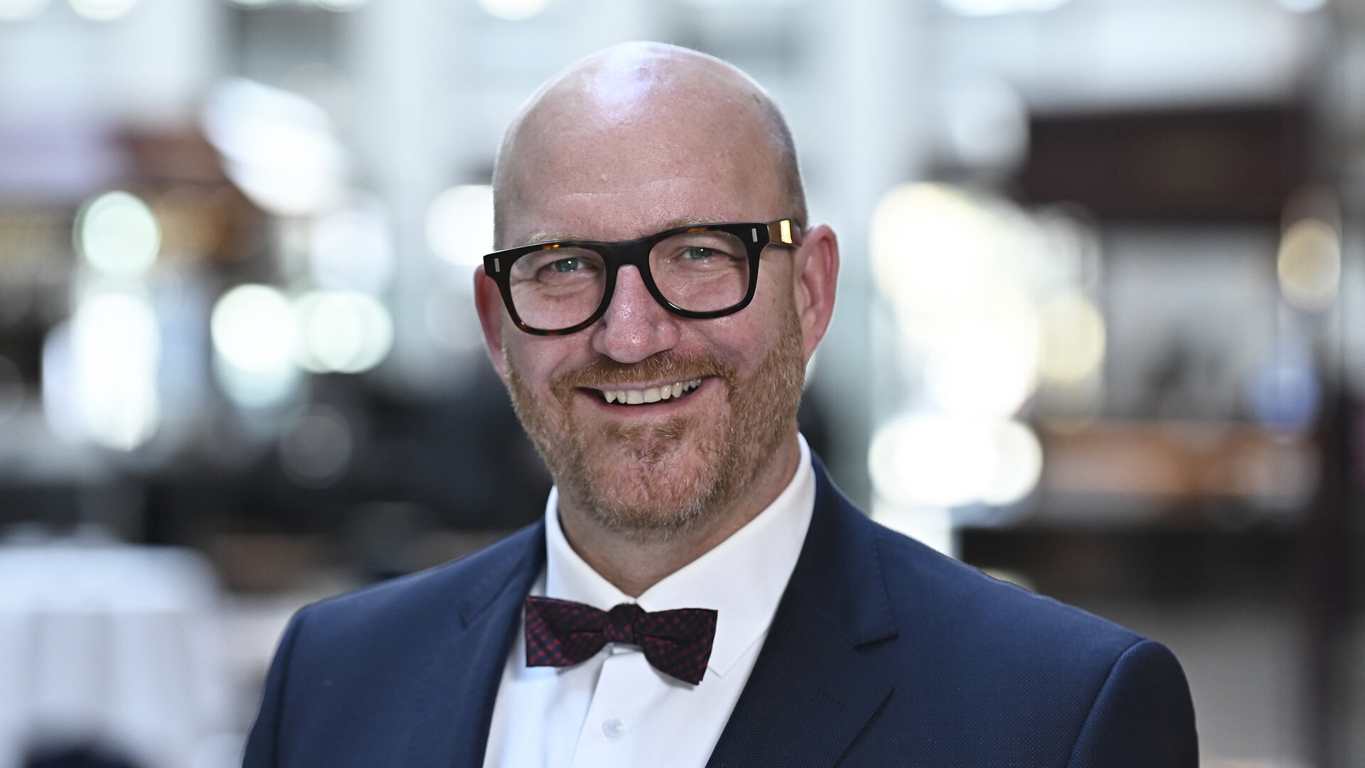 Ein Mann mit Brille und Bart, in einem blauen Anzug mit Fliege, lächelt in die Kamera