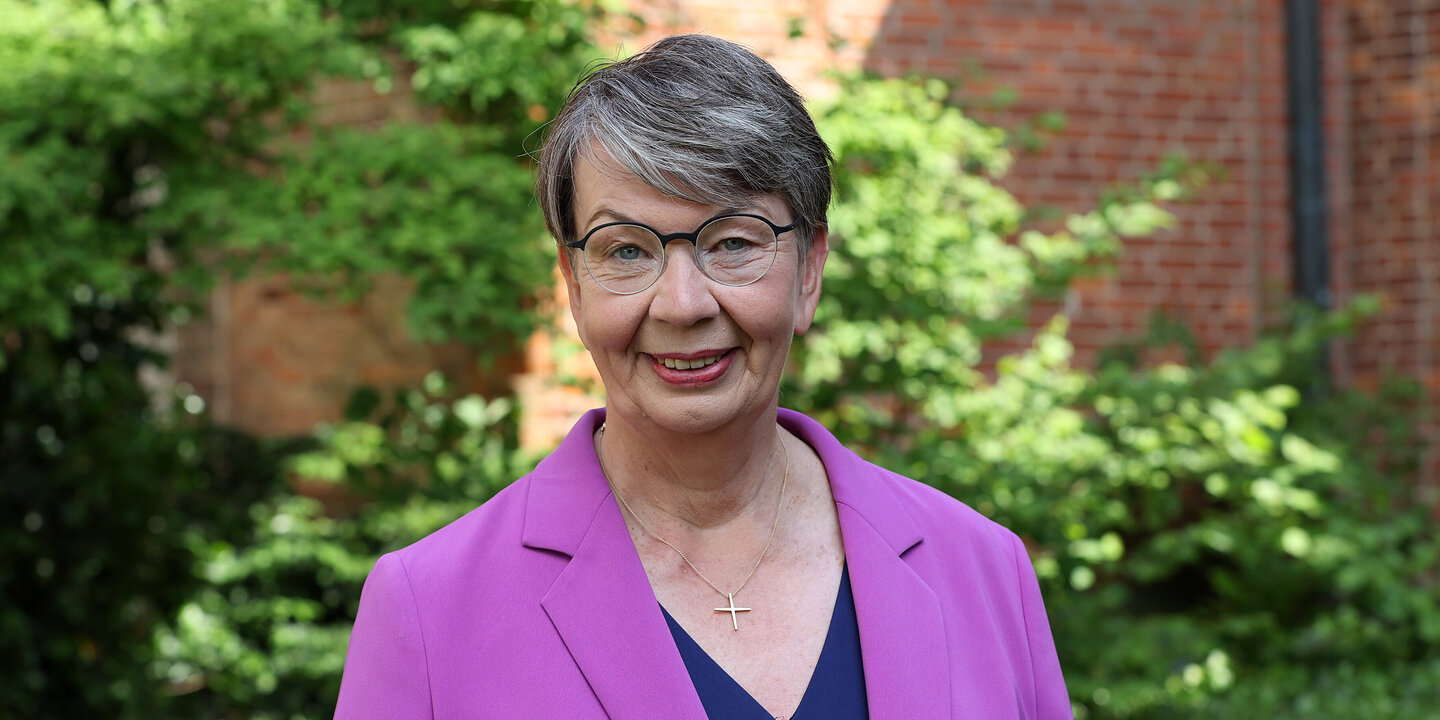 Eine Frau mit Brille und einer violetten Jacke steht vor einem grünen Busch.