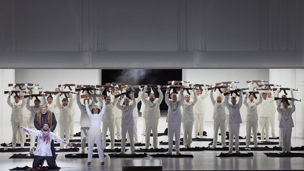 Eine Opernbühne, weißer Hintergrund. Auf der Bühne stehen viele Personen in weißer Kleidung und halten mit beiden Händen Gewehre hoch. Im Vordergrund kniet ein Mann mit blutbeflecktem Hemd; hinter ihm steht ein Mann und würgt ihn.