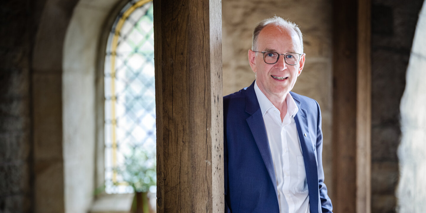 Ein Mann mit Brille, weißem Hemd und blauem Anzug steht in einem alten Klostergebäude und lehnt sich mit der Schulter gegen eine Holzsäule.