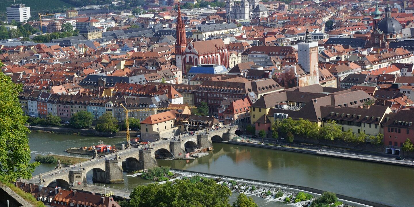 Blick auf eine Stadt im Sommer. Im Vordergrund ein breiter Fluss, über den eine alte Steinbrücke führt.