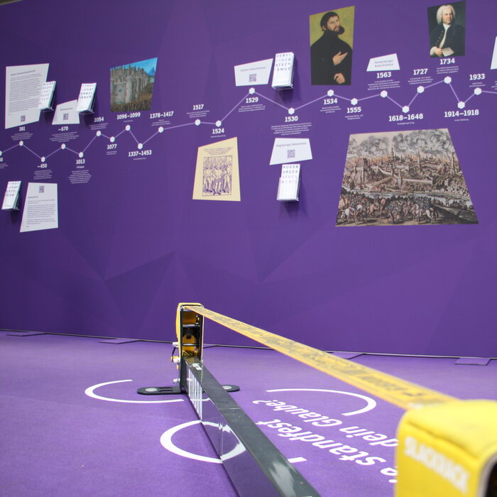 Eine gelbe Slackline vor einer violettem Wand mit einem Zeitstrahl und vielen Bildern