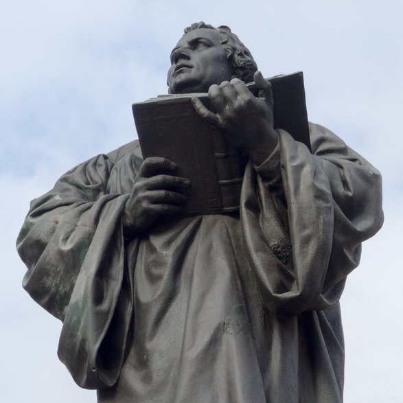 Eine Statue Martin Luthers, der ein offenes Buch hält. Im Hintergrund ein blauer Himmel mit Schleierwolken.