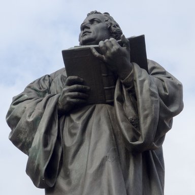 Eine Statue Martin Luthers, der ein offenes Buch hält. Im Hintergrund ein blauer Himmel mit Schleierwolken.