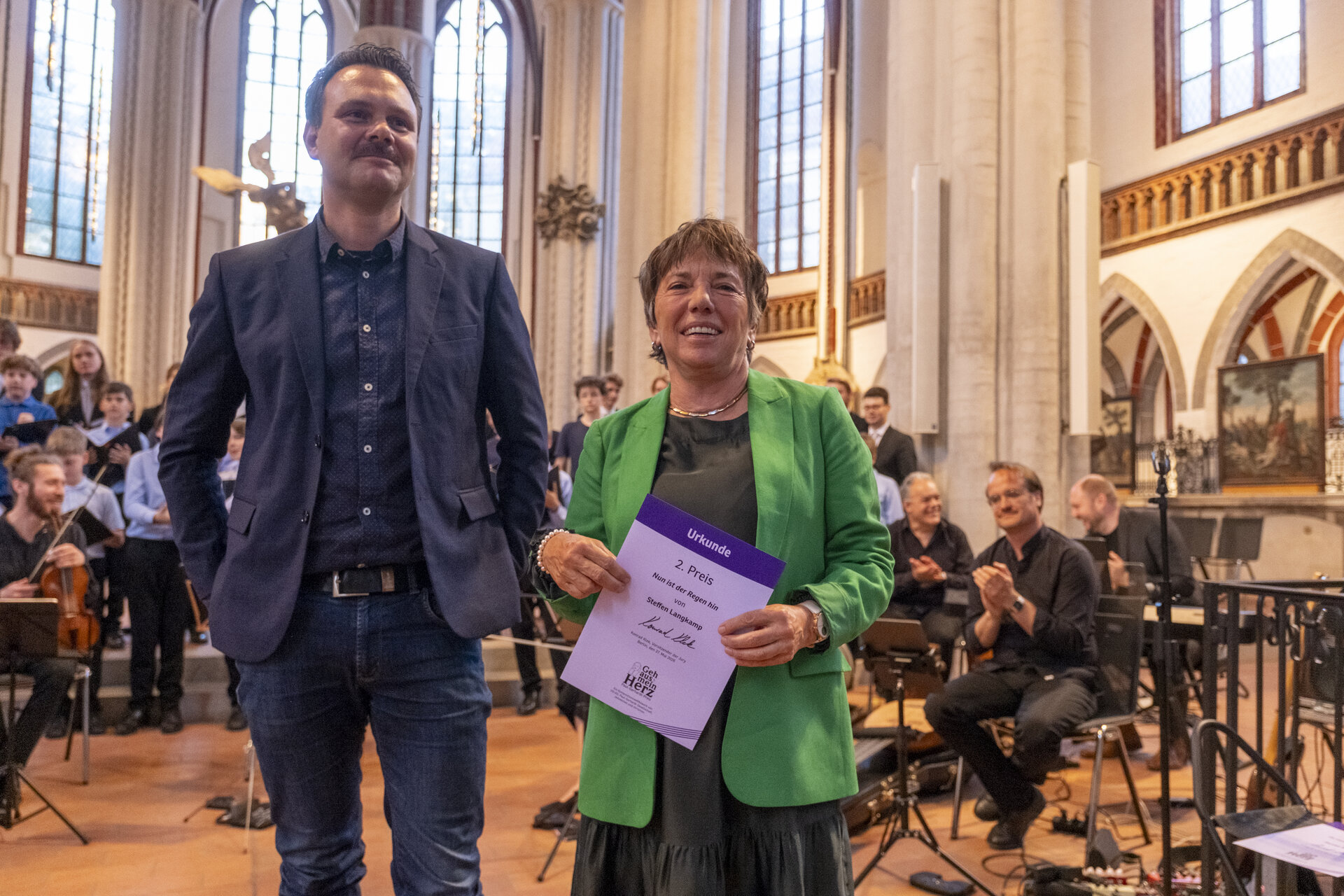 Ein Mann in dunklem Sakko steht neben einer Frau in grünem Blazer.