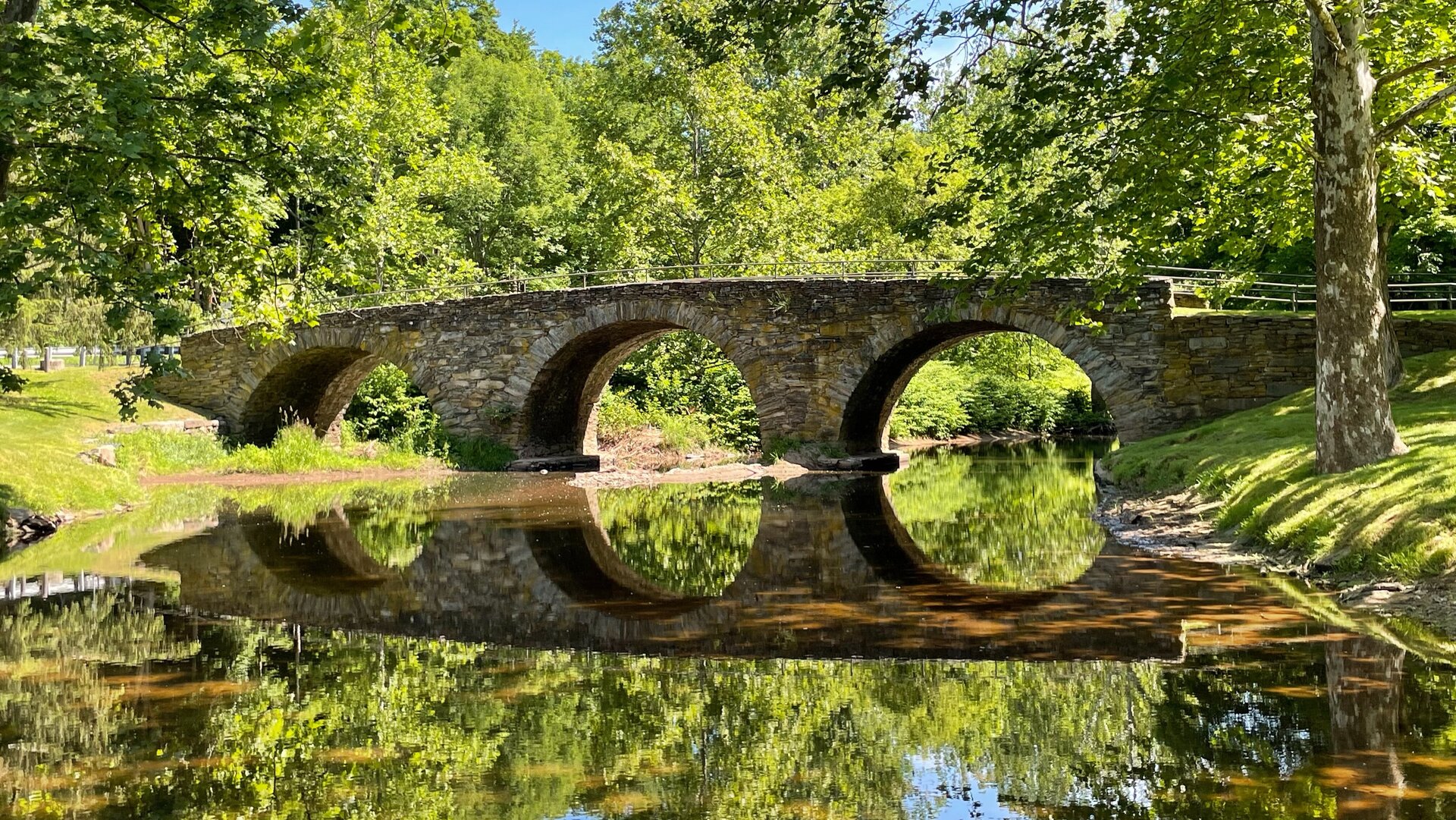 Eine steinerne Brücke spannt sich über einen Fluss. Die Ufer sind mit Gras und Bäumen bewachsen. Im Fluss spiegelt sich die Brücke.