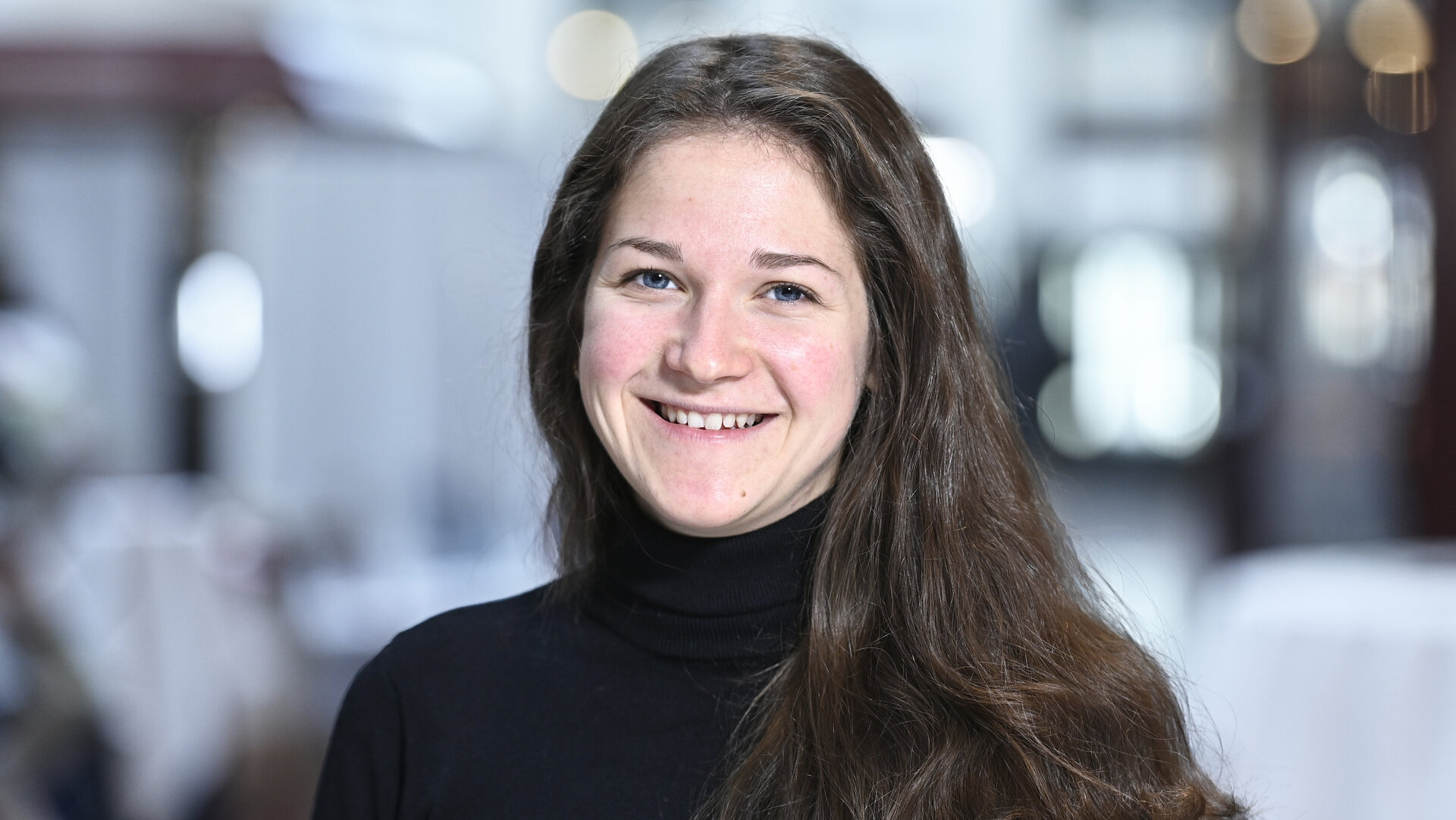 Eine junge Frau mit langen braunen Haaren und einem schwarzen Rollkragenpullover lächelt in die Kamera