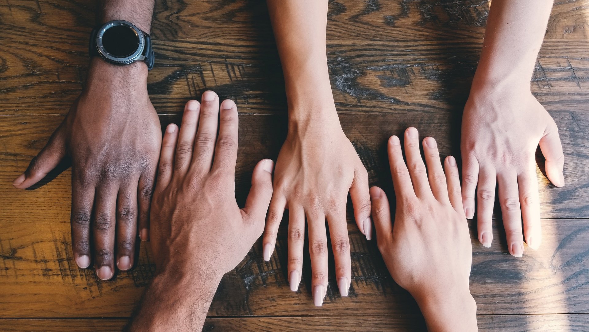 fünf Hände verschiedener Hautfarben liegen auf einem Holztisch