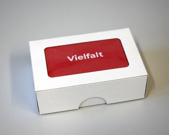 Eine weiße Pappschachtel, in der eine rote Karte mit der Aufschrift "Vielfalt" liegt.