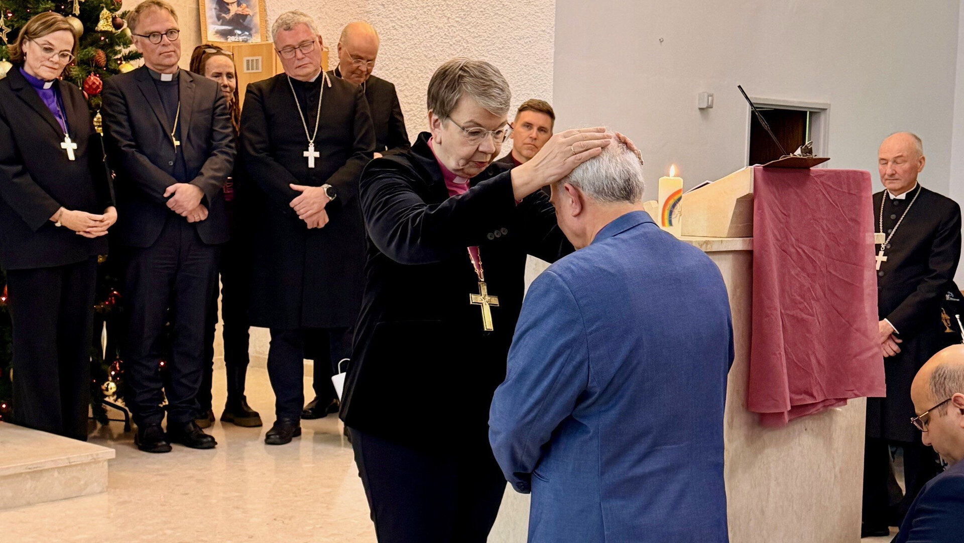 Kirchliche Segnung, bei der eine Geistliche einem Mann die Hände auf den Kopf legt, umgeben von weiteren Geistlichen in einem Kirchenraum