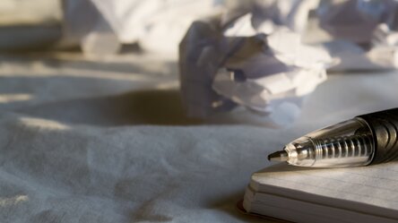 Mehrere zu Bällen zerknüllte Blätter Papier liegen auf einem Tisch. Im Vordergrund ist ein Block und ein darauf liegender Kugelschreiber zu erkennen.
