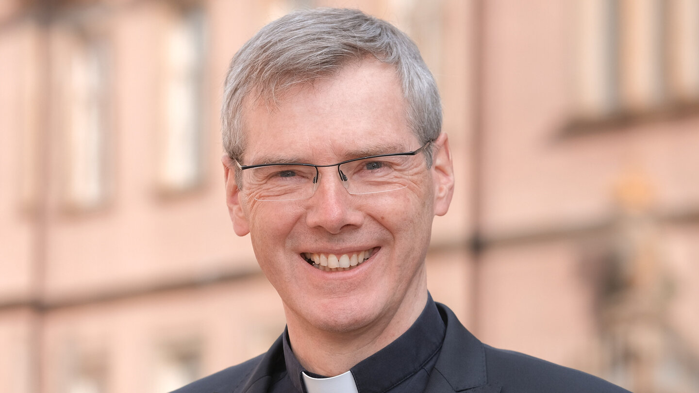 Lächelnder Mann mit grauen Haaren und Brille im schwarzen Anzug mit Priesterkragen, unscharfe Gebäudefassade im Hintergrund.