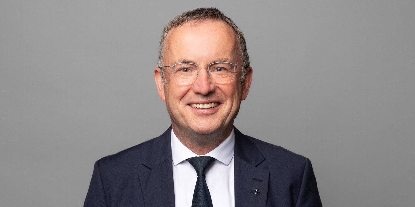 Ein bartloser Mann mit Brille in dunklem Anzug, weißem Hemd und Krawatte lächelt in die Kamera.